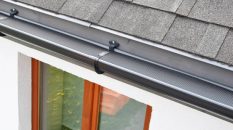 Plastic guard over new dark grey plastic rain gutter on asphalt shingles roof and white color decorative plaster facade with brown wooden window with reflection on glass pane.
